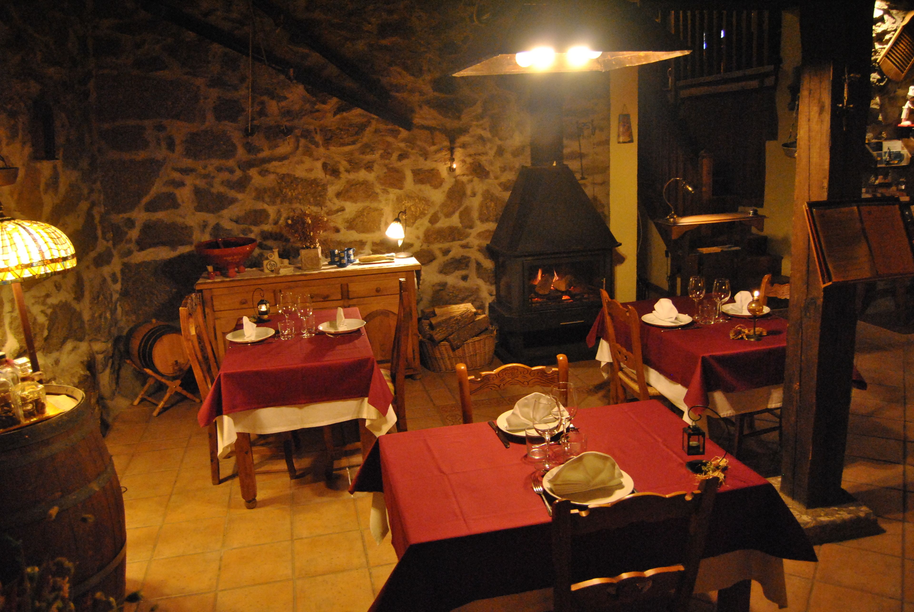 Zona de menjador rústica amb parets de pedra, mobles de fusta i una llar de foc. Taules preparades amb mantells vermells i vaixella blanca.