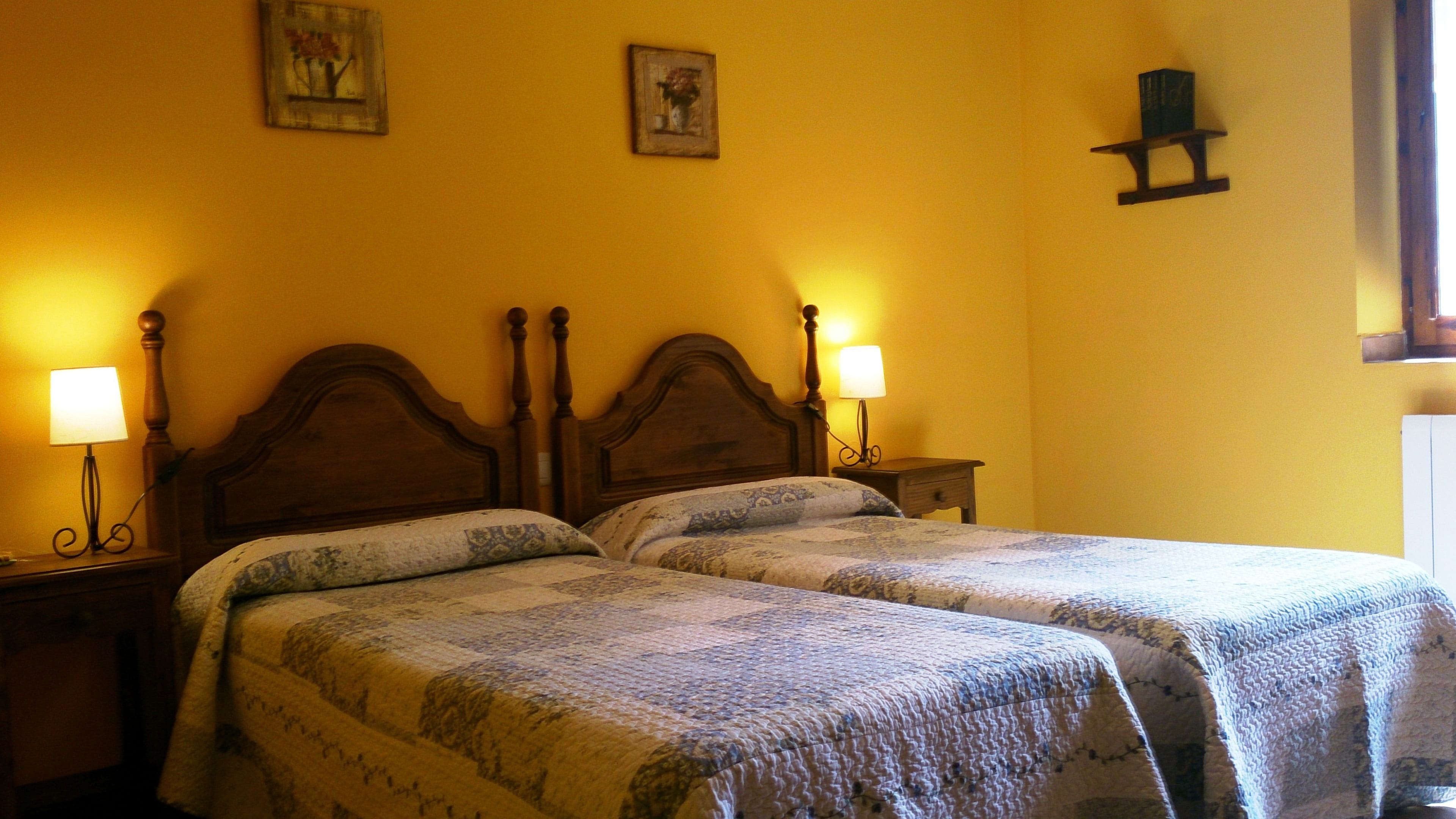 A room with two beds, each with a wooden headboard and a bedside table with a lamp.