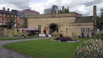 A historic building with a stone facade, an arched entrance, and a garden with manicured lawns and flower beds.