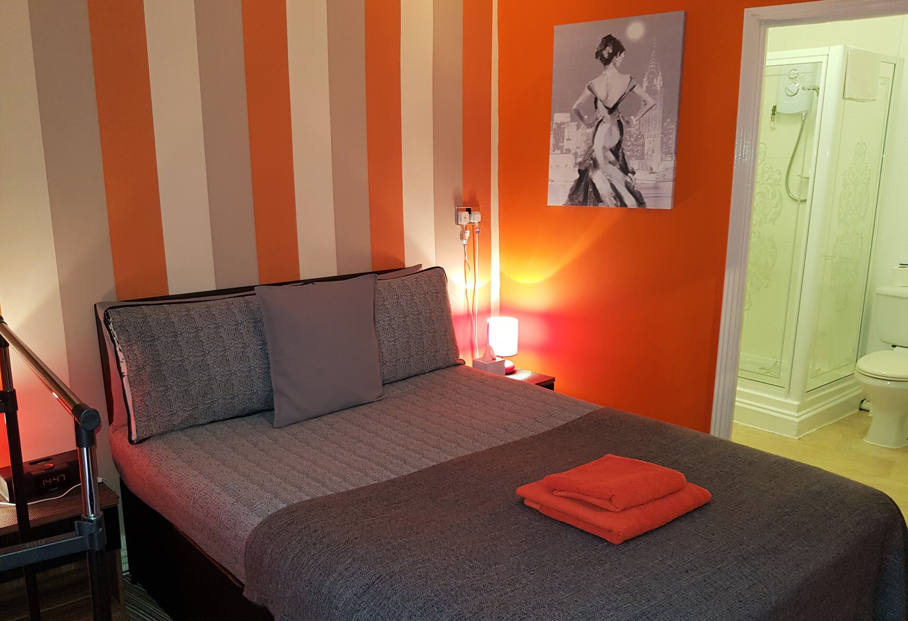 Habitación de hotel con cama, mesita de noche con lámpara, baño con ducha e imagen en la pared.