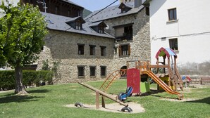 Un parque infantil con un balancín, tobogán y estructura de escalada frente a un edificio de piedra con techo inclinado.