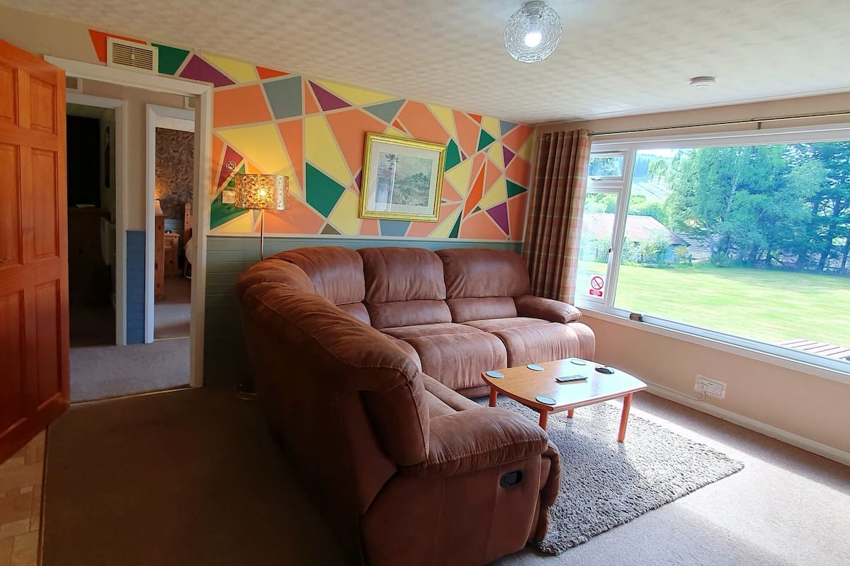 A living room with a brown leather sofa, a small wooden coffee table, and a large window offering a view of a green lawn.