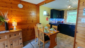 A wooden interior with a dining table set for two, a clock on the wall, and a kitchen area with a window. - Torcroft Lodges (Inverness)