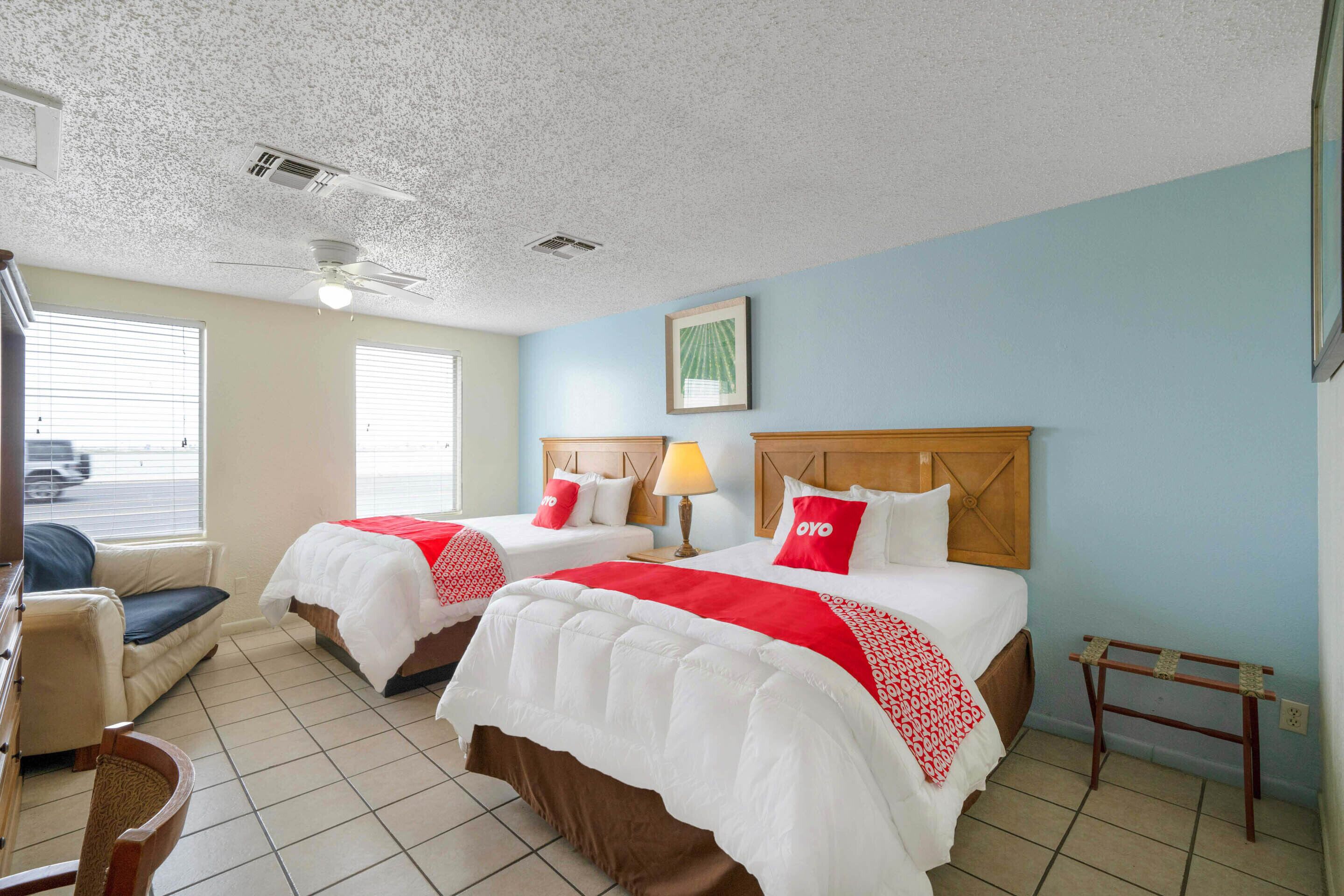 A hotel room with two beds, a ceiling fan, a window with blinds, a chair, and a small table.