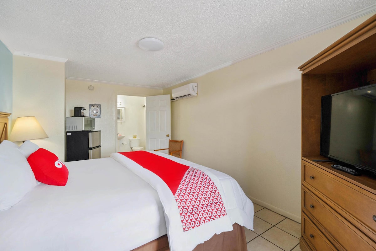 Una habitación de hotel con cama, televisor, cómoda, escritorio con lámpara y kitchenette.