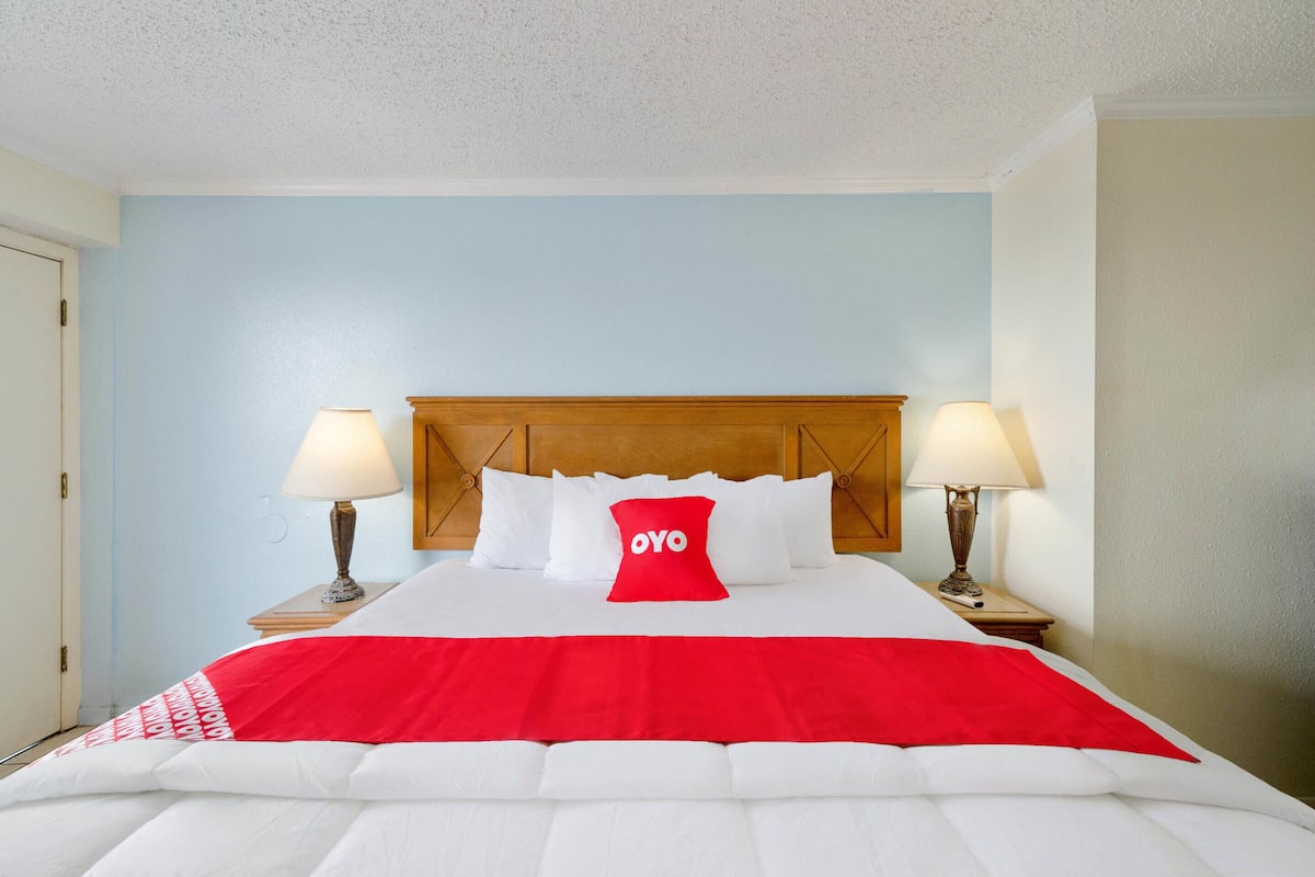 Habitación de hotel con cama, dos lámparas de noche, cabecera de madera y almohada roja con la palabra 'OYO'.