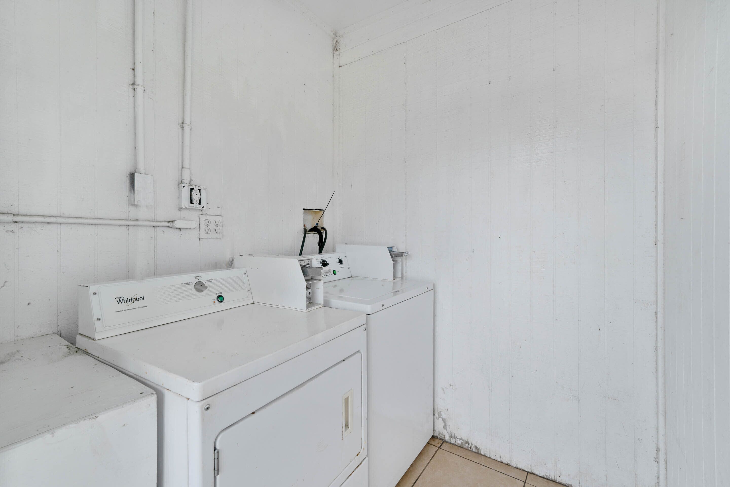 Une buanderie équipée de deux machines à laver et de deux sèche-linge Whirlpool blancs, d’un sol carrelé et d’un mur blanc où l’on peut voir des tuyaux et des prises de courant.