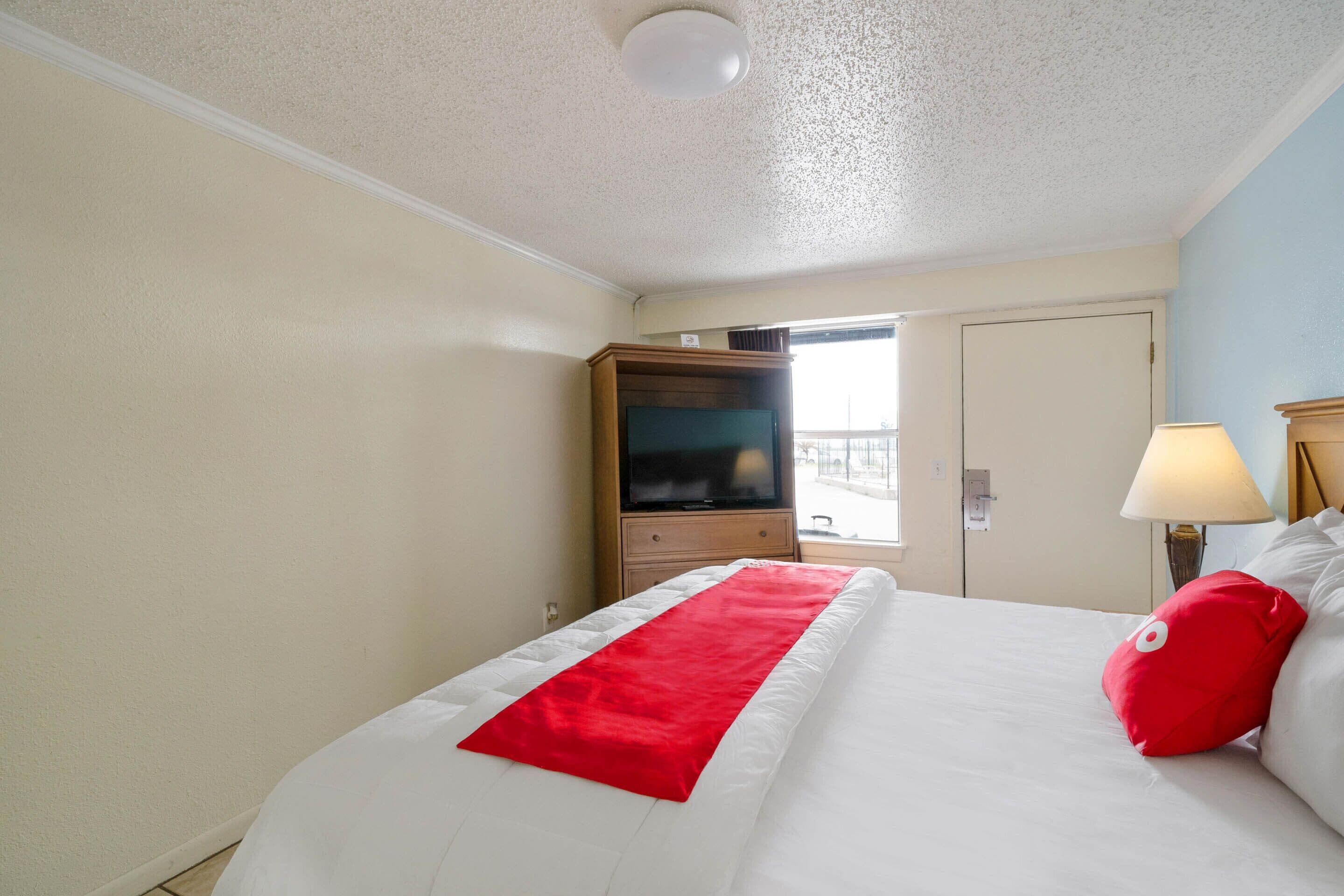 Une chambre d’hôtel avec un lit, un plaid rouge, un coussin rouge, une télévision et une lampe.