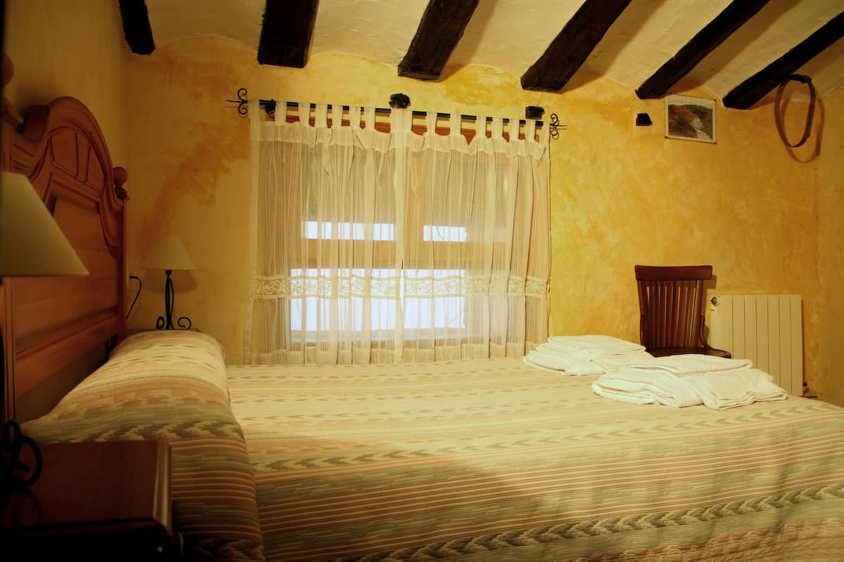 Un dormitorio con cama, mesita de noche, lámpara, ventana con cortinas translúcidas y cabecero de madera.