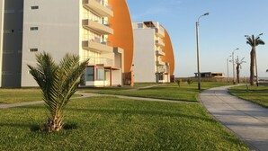 A modern building with multiple balconies, a curved orange facade, and a well-maintained lawn with palm trees. - Apartamento Frente Islas Ballestas (Paracas)