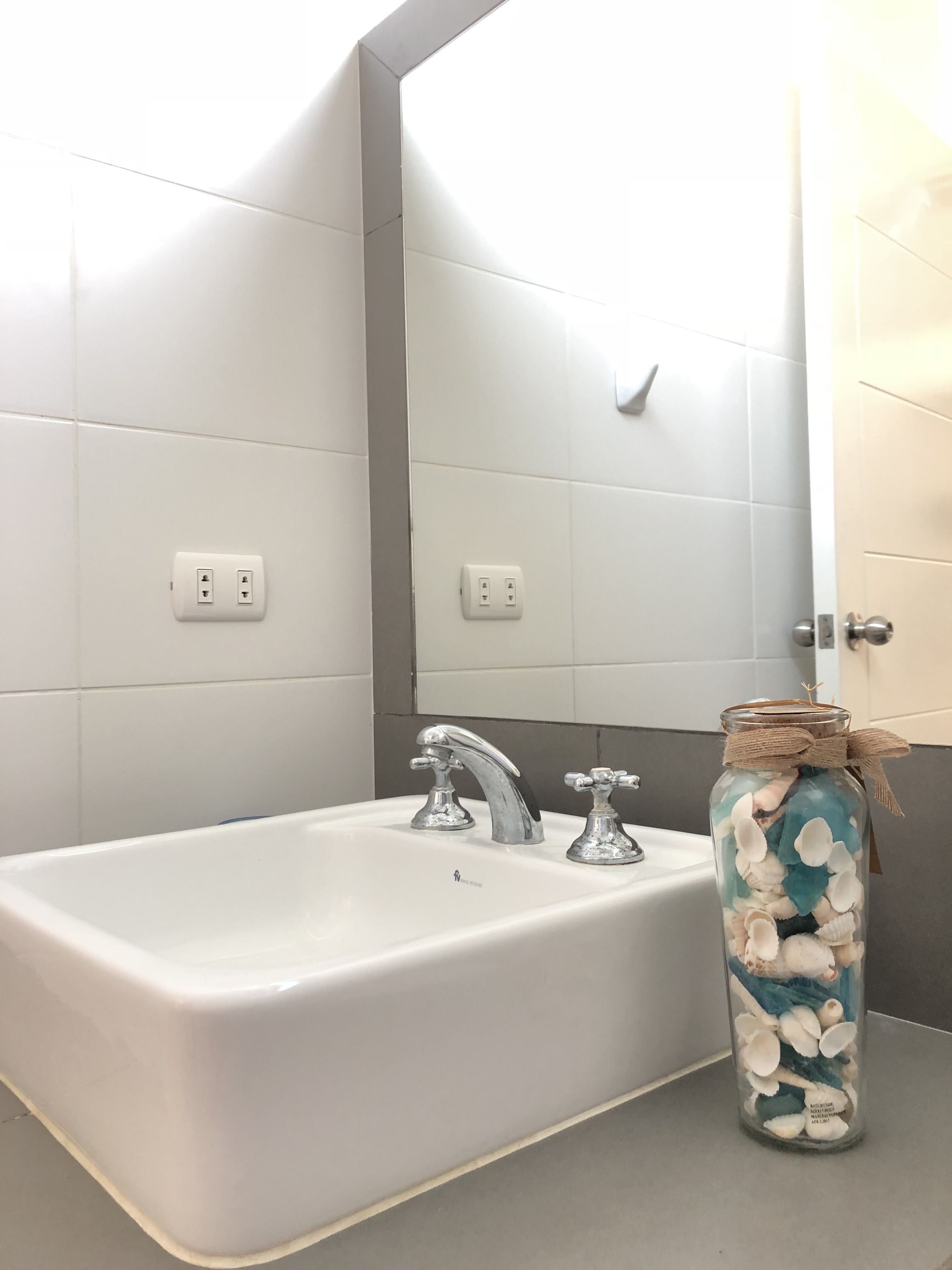 Ein Badezimmer mit einem weißen Waschbecken, zwei Wasserhähnen und einem Glas mit Meeresschnecken auf der Ablagefläche.