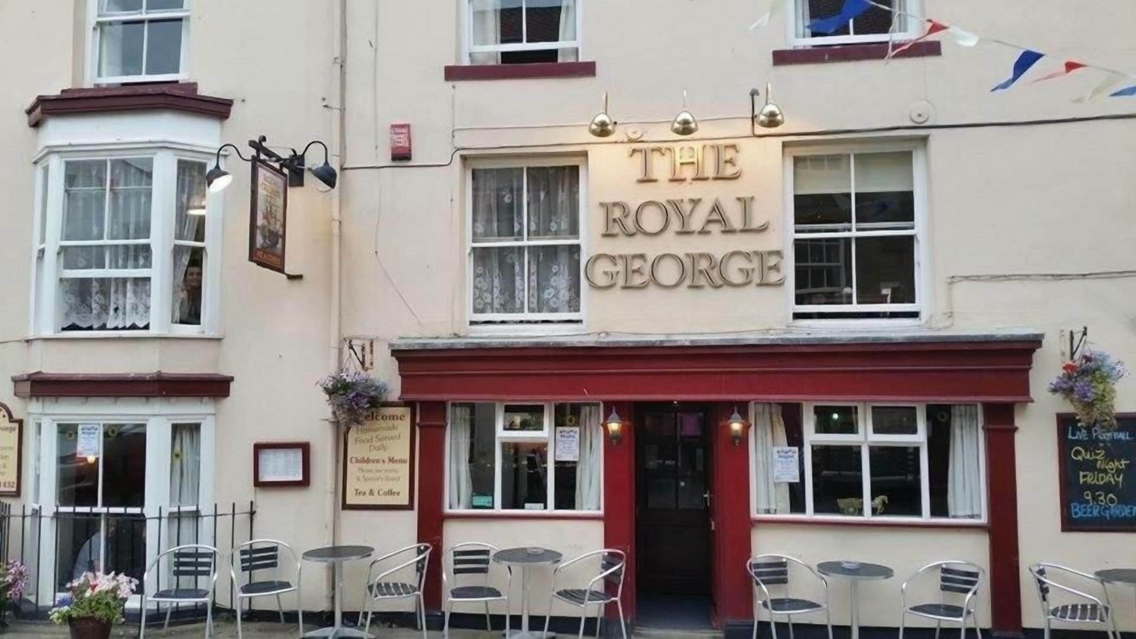 Un bâtiment sur lequel on peut lire l’inscription « The Royal George », avec une terrasse extérieure dotée de chaises et de tables en métal.