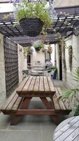 Una mesa de picnic de madera con bancos, rodeada por una pérgola con plantas colgantes, en un patio con una reja de metal y suelo de hormigón.