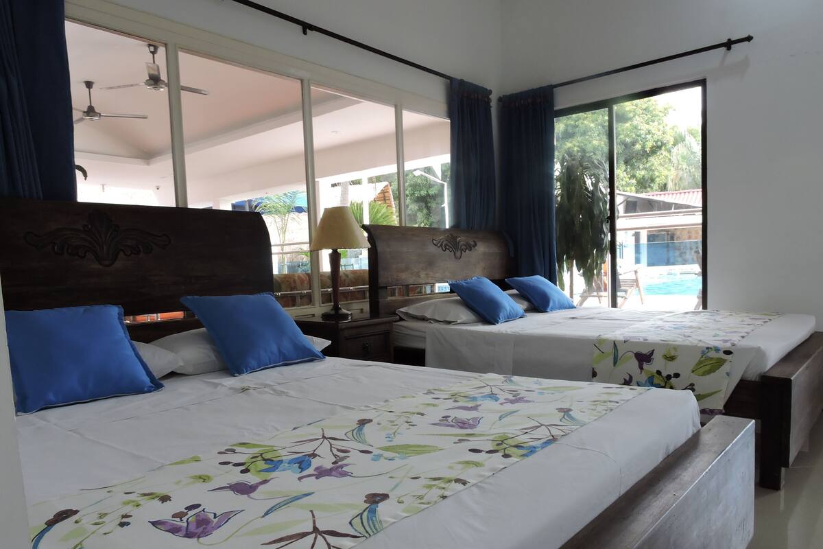 Habitació d'hotel amb dos llits, una finestra gran amb cortines, un ventilador de sostre i vista a una piscina i vegetació a l'exterior.