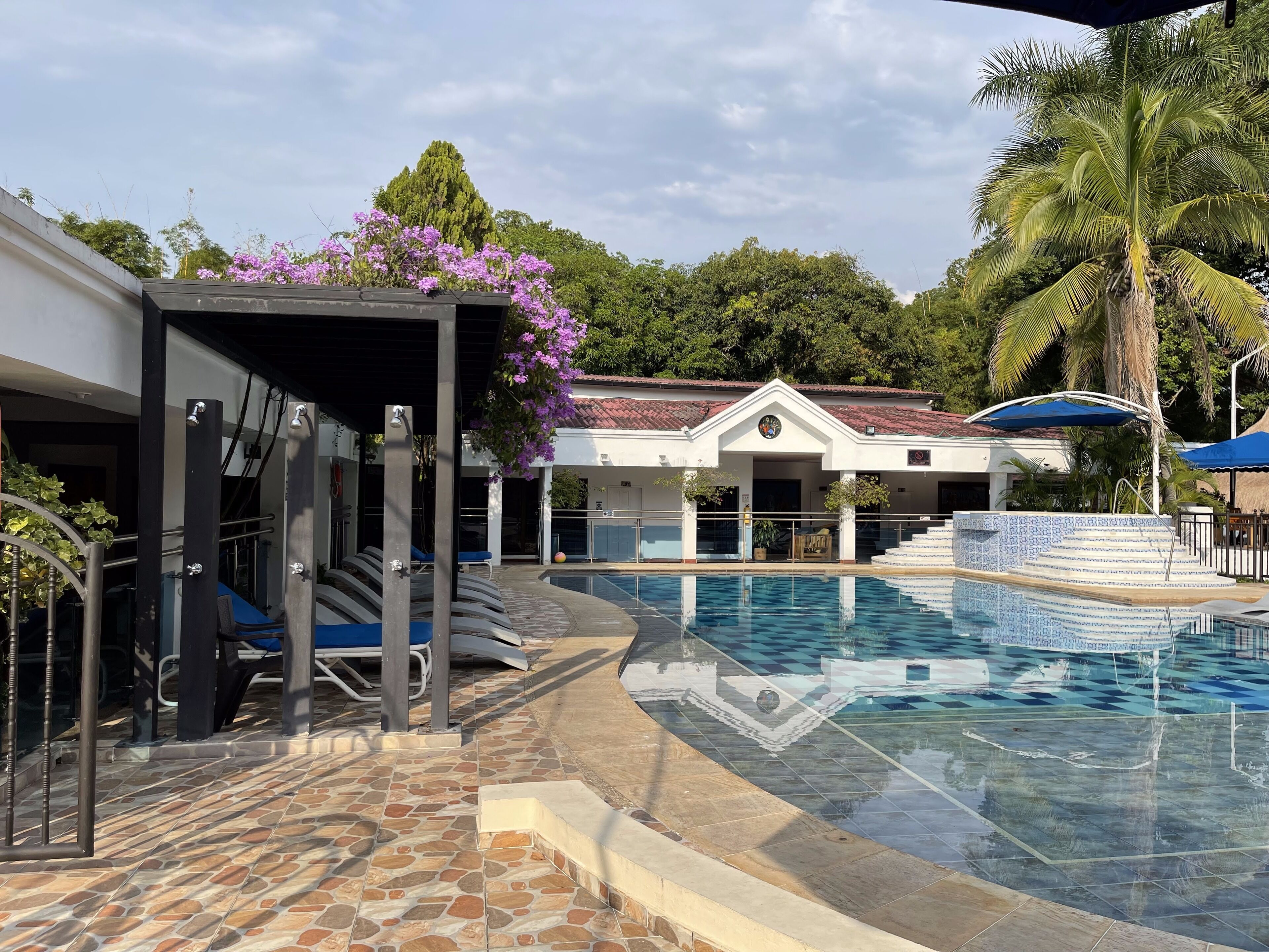 A swimming pool with a stone-tiled deck, a covered area with exercise equipment, and a building with a red-tiled roof.
