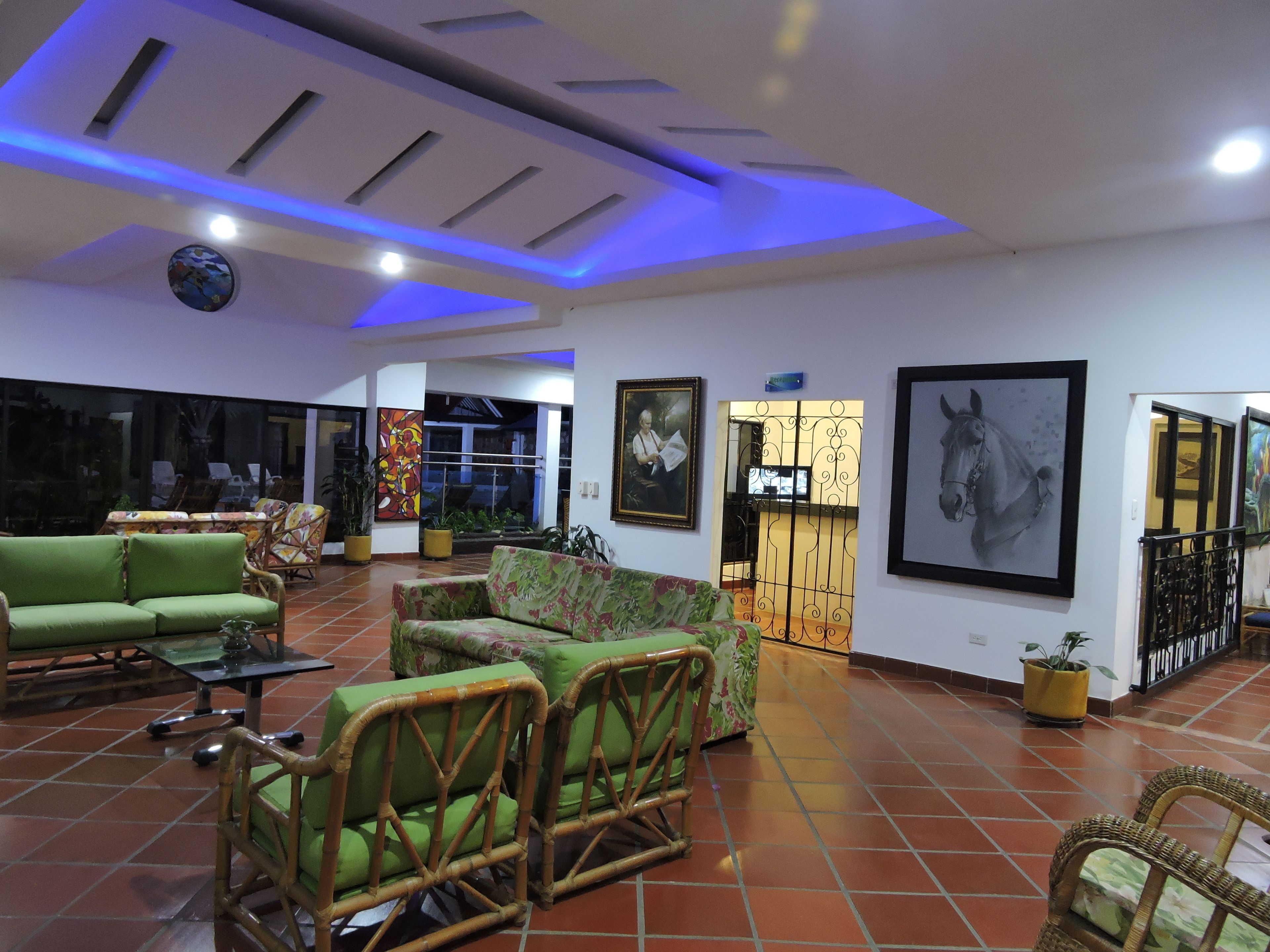Uma sala ampla com sofás verdes e cadeiras de vime, piso em azulejos e obras de arte nas paredes.
