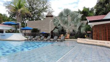 Ein Swimmingpool mit Liegestühlen, ein Gebäude mit Reetdach und Palmen.
