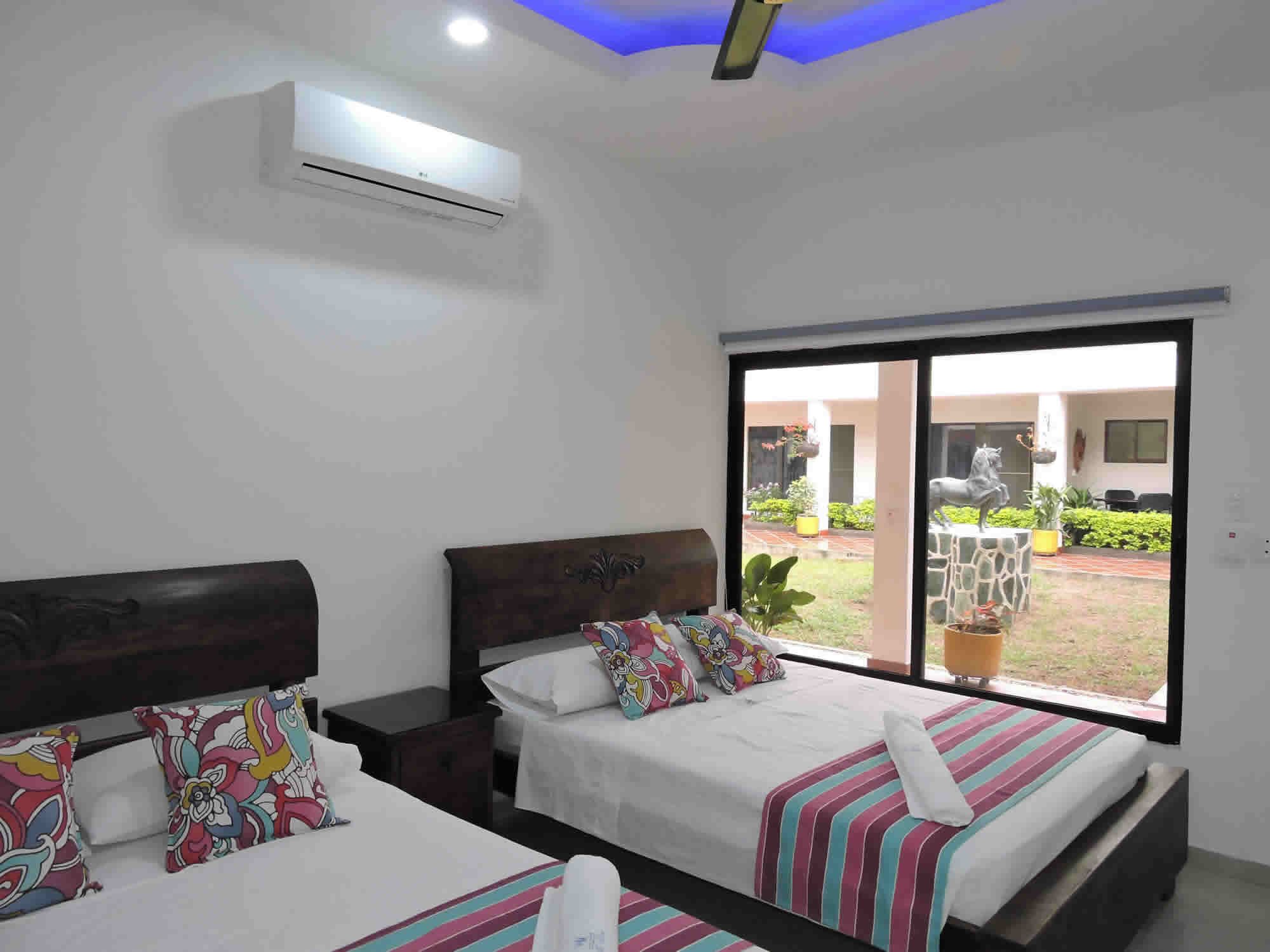 Habitación de hotel con dos camas, almohadas coloridas, un ventanal grande y un equipo de aire acondicionado.