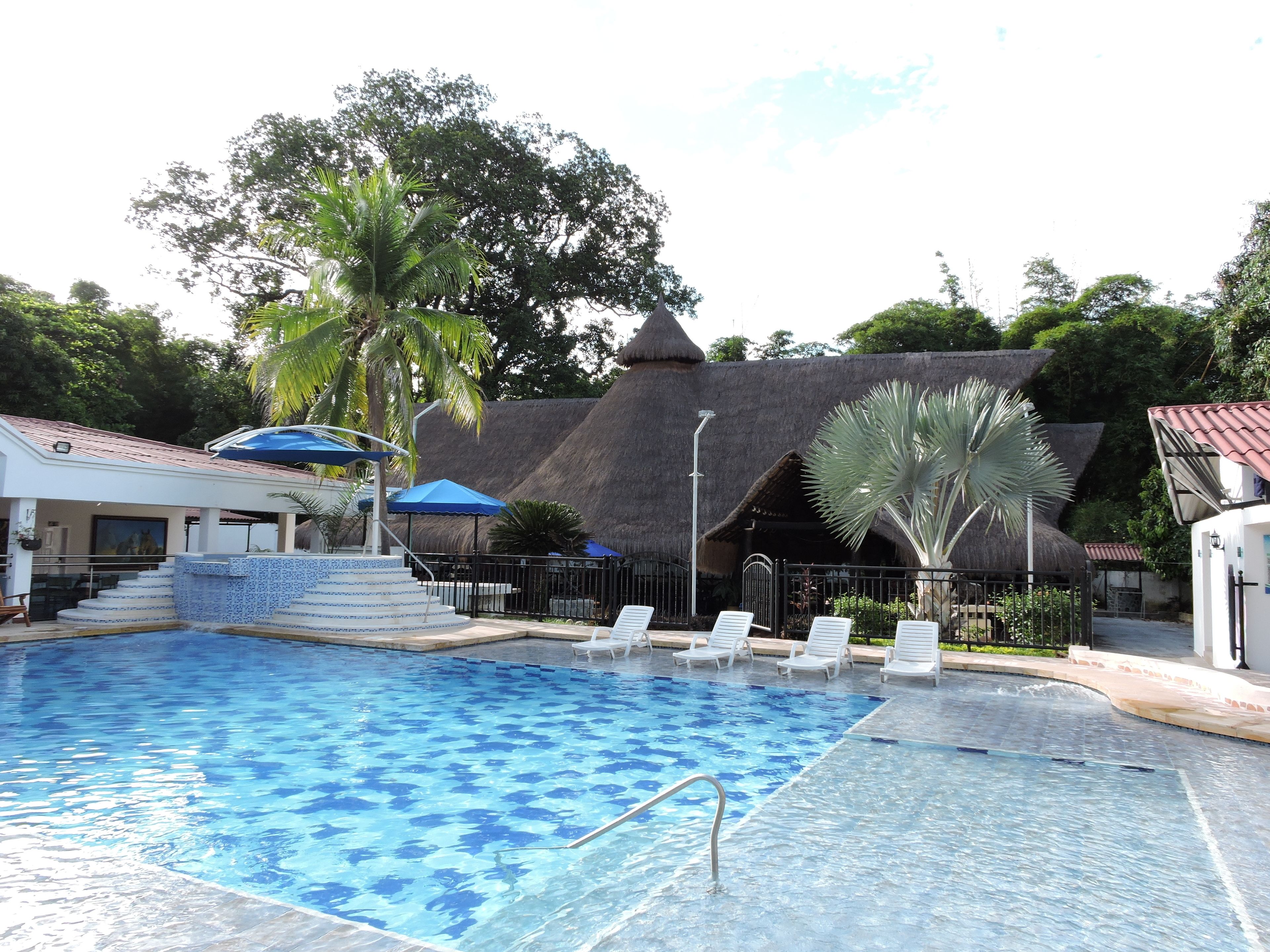 Una piscina con un patrón de azulejos azules, rodeada de sillas reclinables blancas y un edificio con techo de paja.