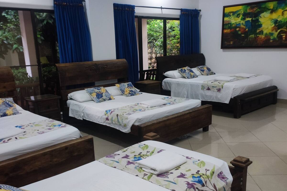 A hotel room with four wooden beds, each with a white bedspread and floral-patterned pillows. There is a ceiling fan, a large window with blue curtains, and a painting on the wall.