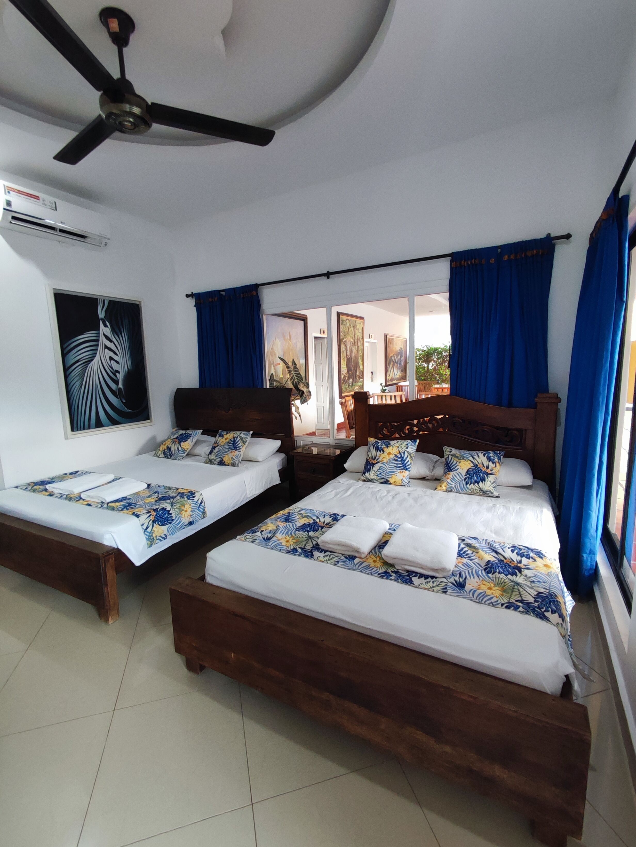 A hotel room with two wooden beds, a ceiling fan, and a large window with blue curtains.