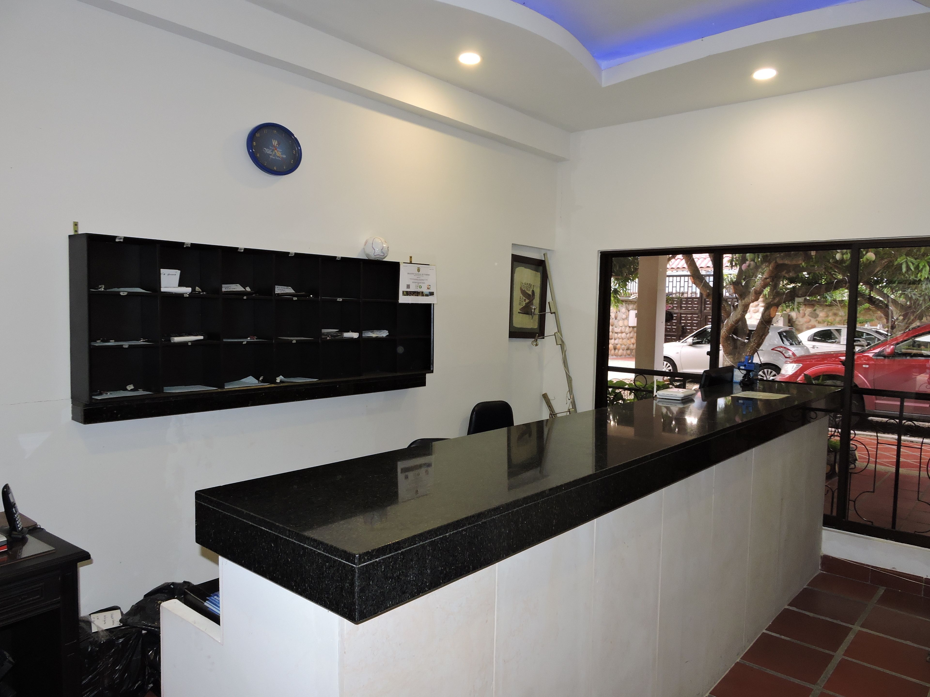 Zona de recepción con mostrador negro, paredes blancas, un reloj y una ventana con vista a coches aparcados.