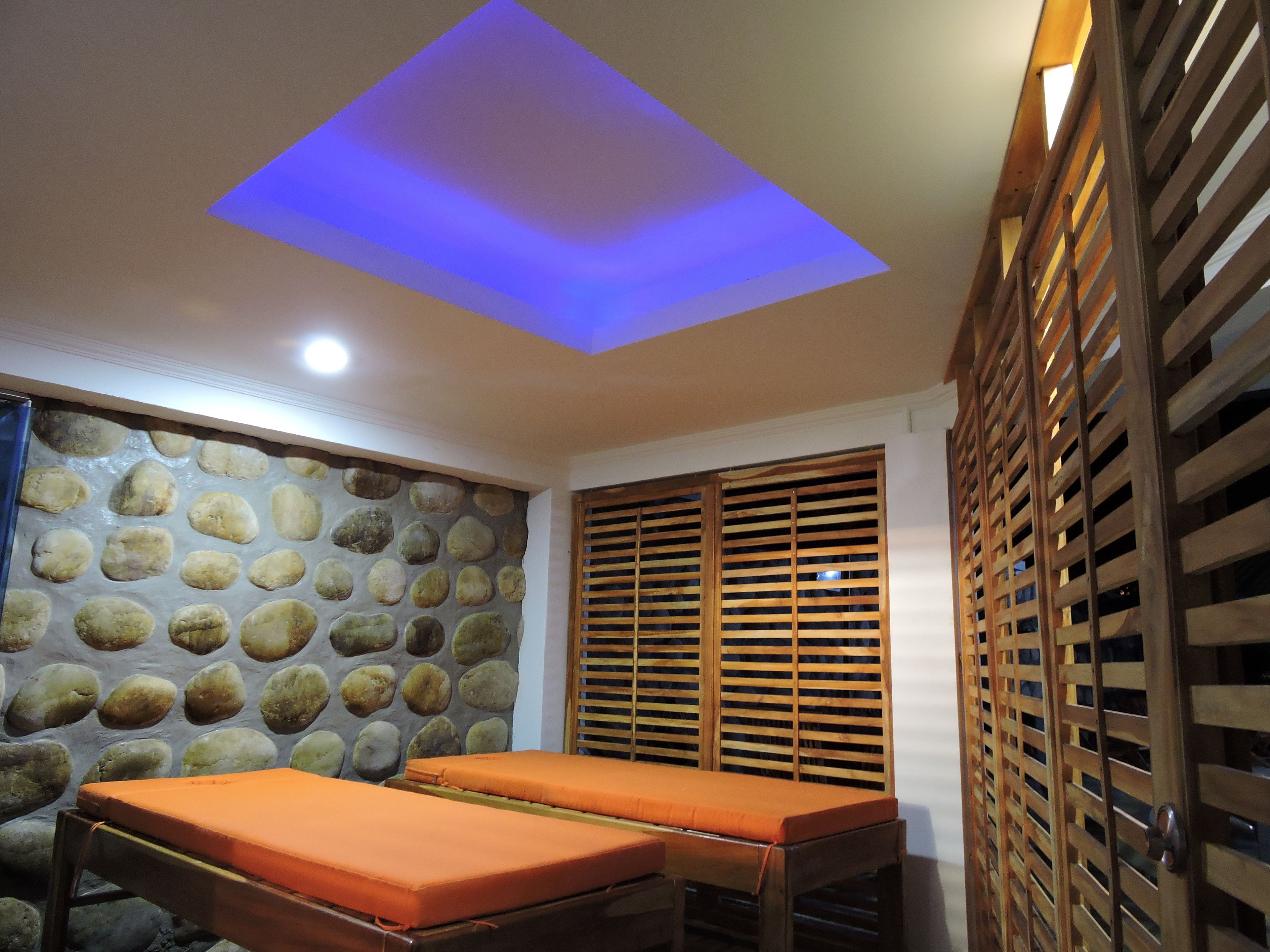 A room with a stone wall, wooden slatted window, and a bed with an orange mattress.