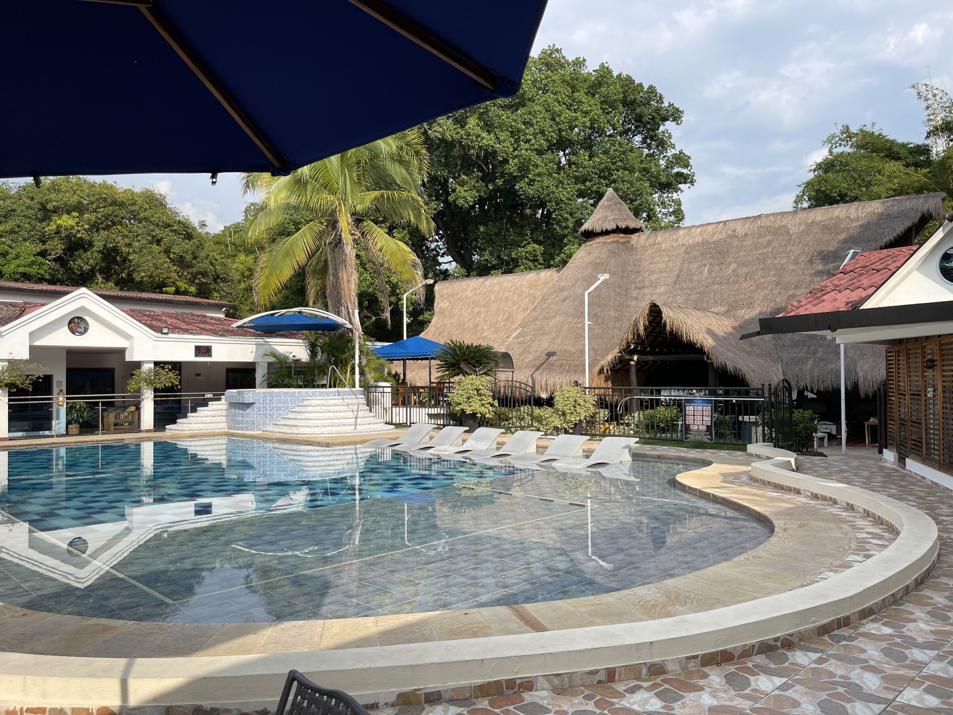A swimming pool with a tiled deck, surrounded by thatched-roof structures and palm trees.