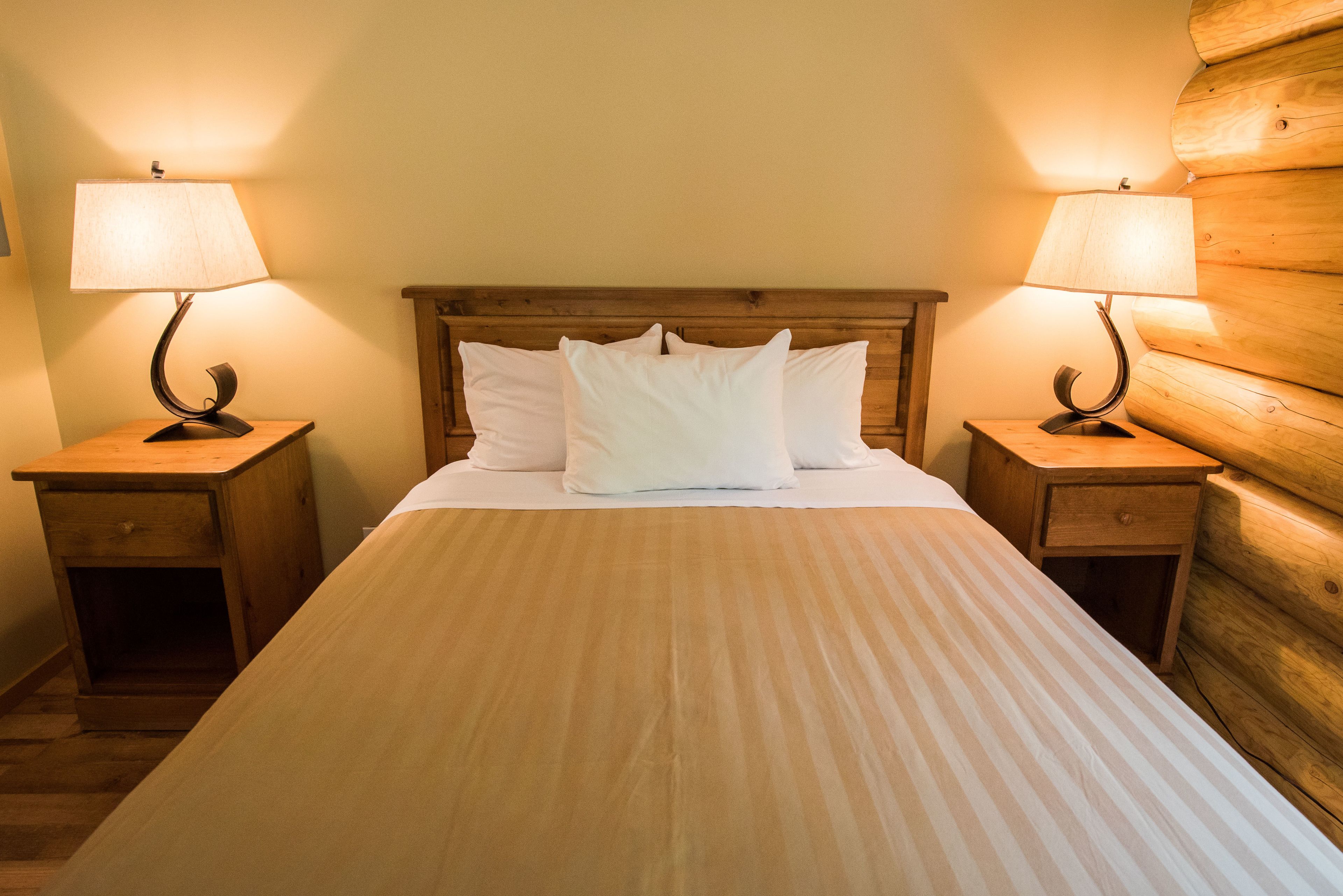 A neatly made bed with a striped bedspread, flanked by two wooden nightstands with lamps, in a room with wooden walls.