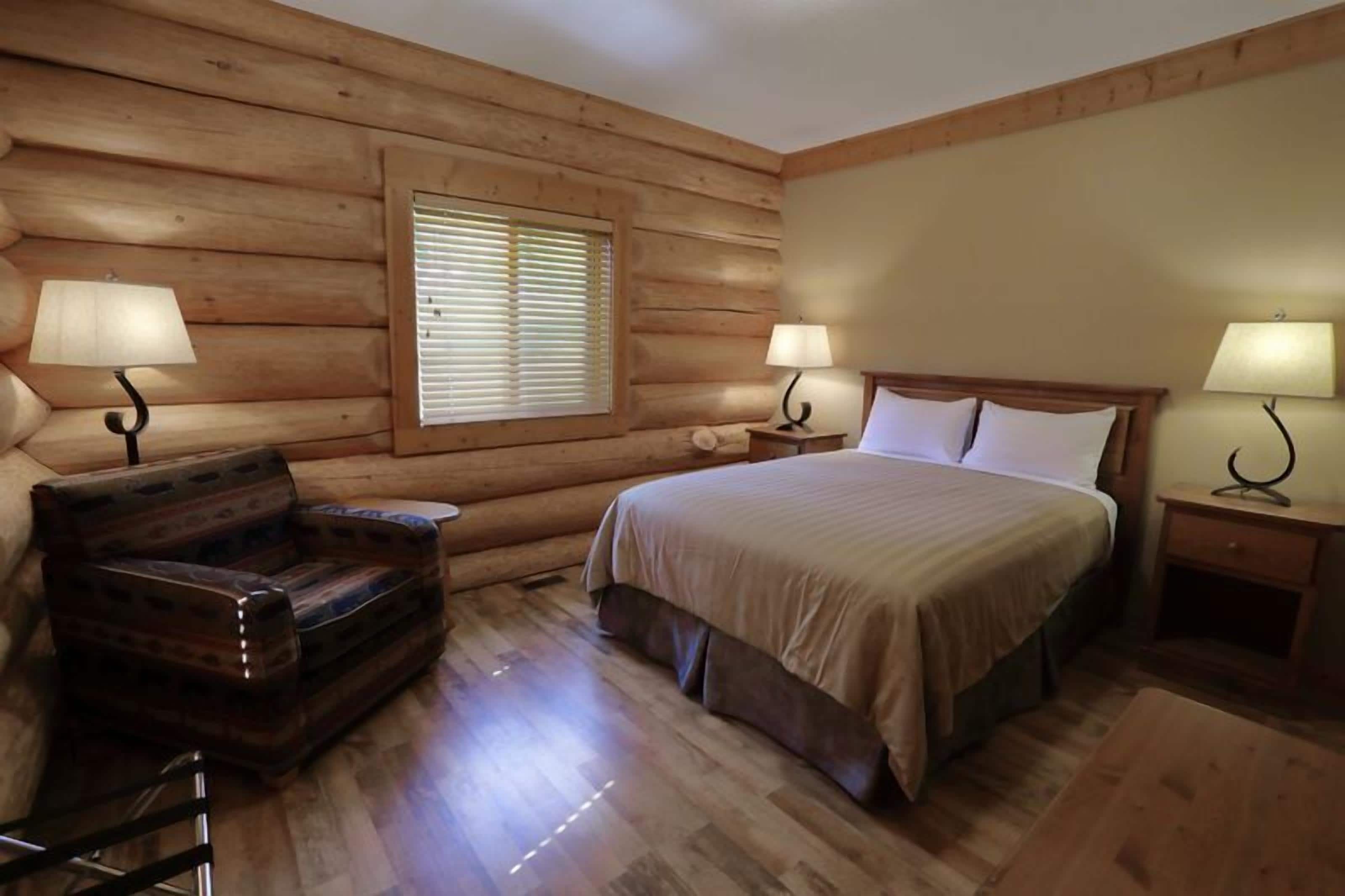 A log cabin bedroom with a bed, two nightstands, two lamps, a wooden chair, and a window with blinds.
