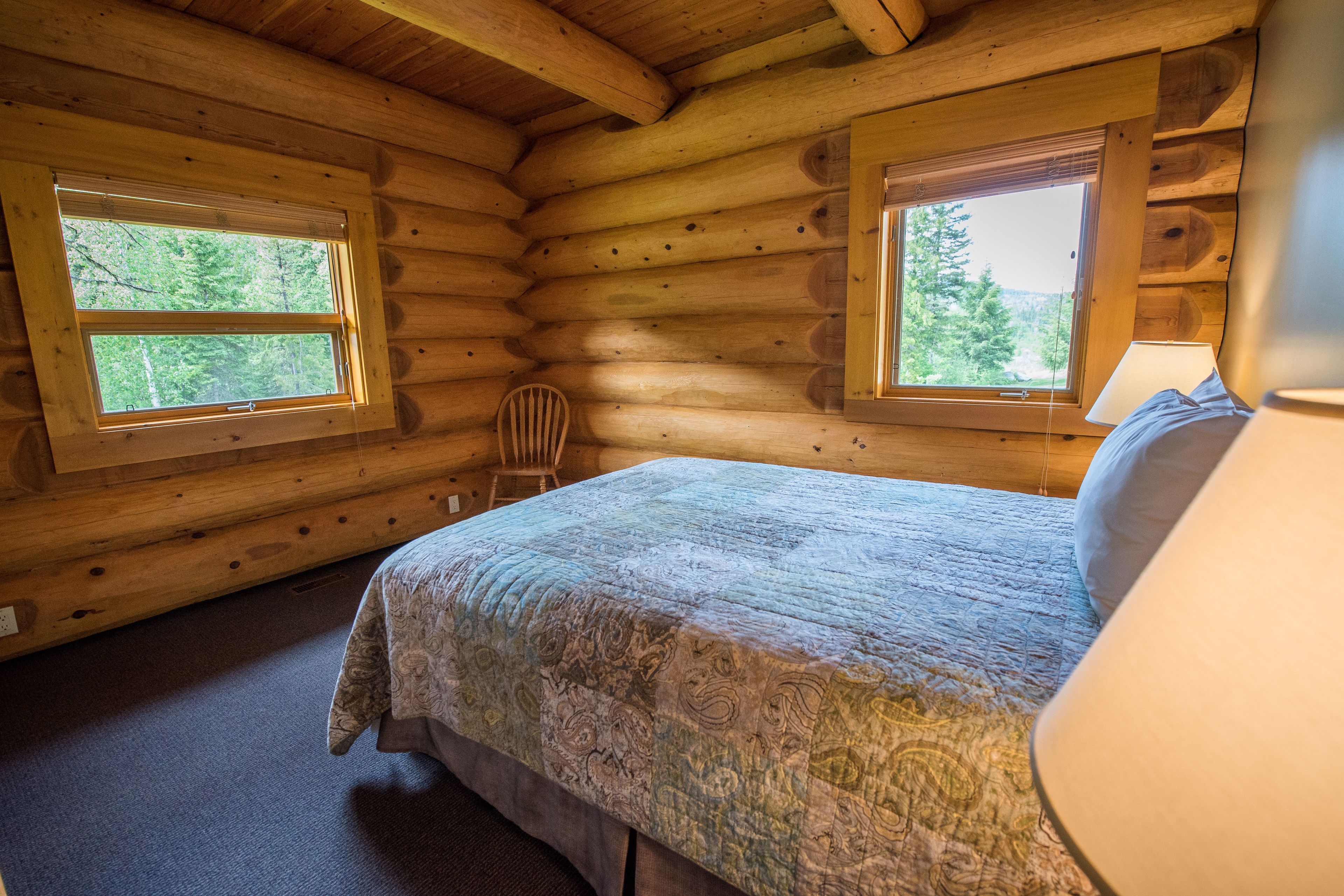 A log cabin bedroom with two windows, a bed, a chair, and a lamp.
