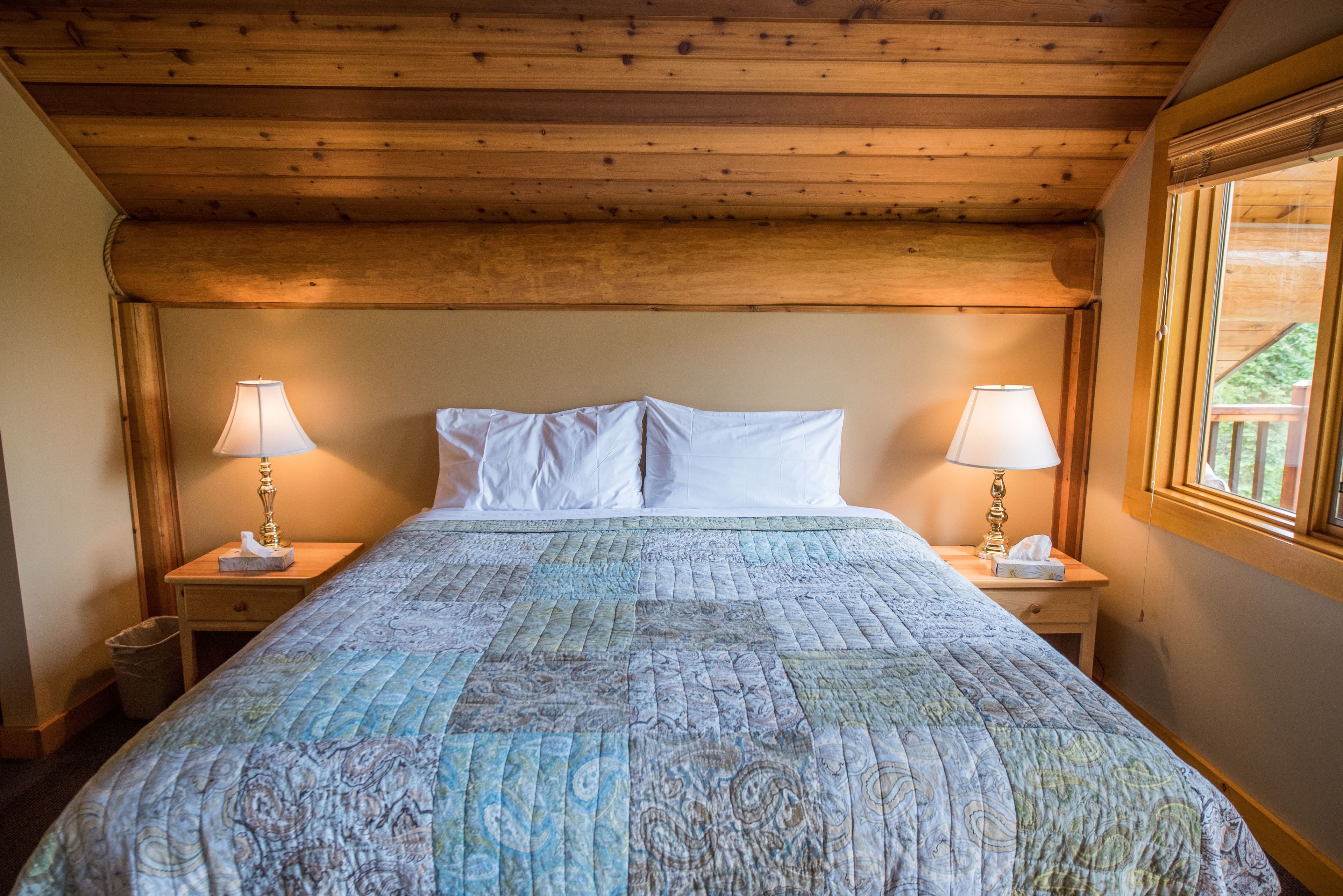 Un dormitorio con techo de madera, colcha acolchada, dos mesitas de noche con lámparas y ventana con vistas al verdor.