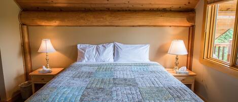 Un dormitorio con techo de madera, colcha acolchada, dos mesitas de noche con lámparas y ventana con vistas al verdor.