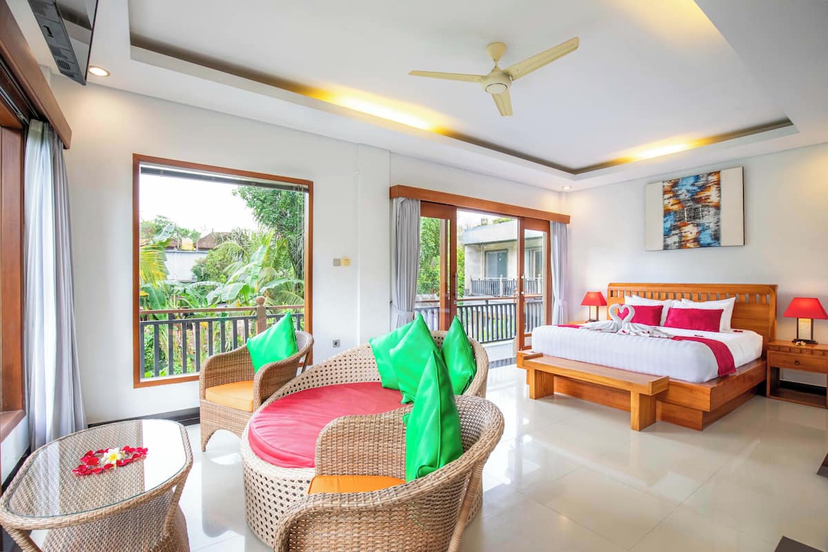 A hotel room with a balcony, wicker furniture, a bed with red pillows, and a ceiling fan.