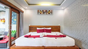 A hotel room with a large bed, wooden headboard, and a view of a balcony with outdoor furniture. - Amoya Inn (Ubud)