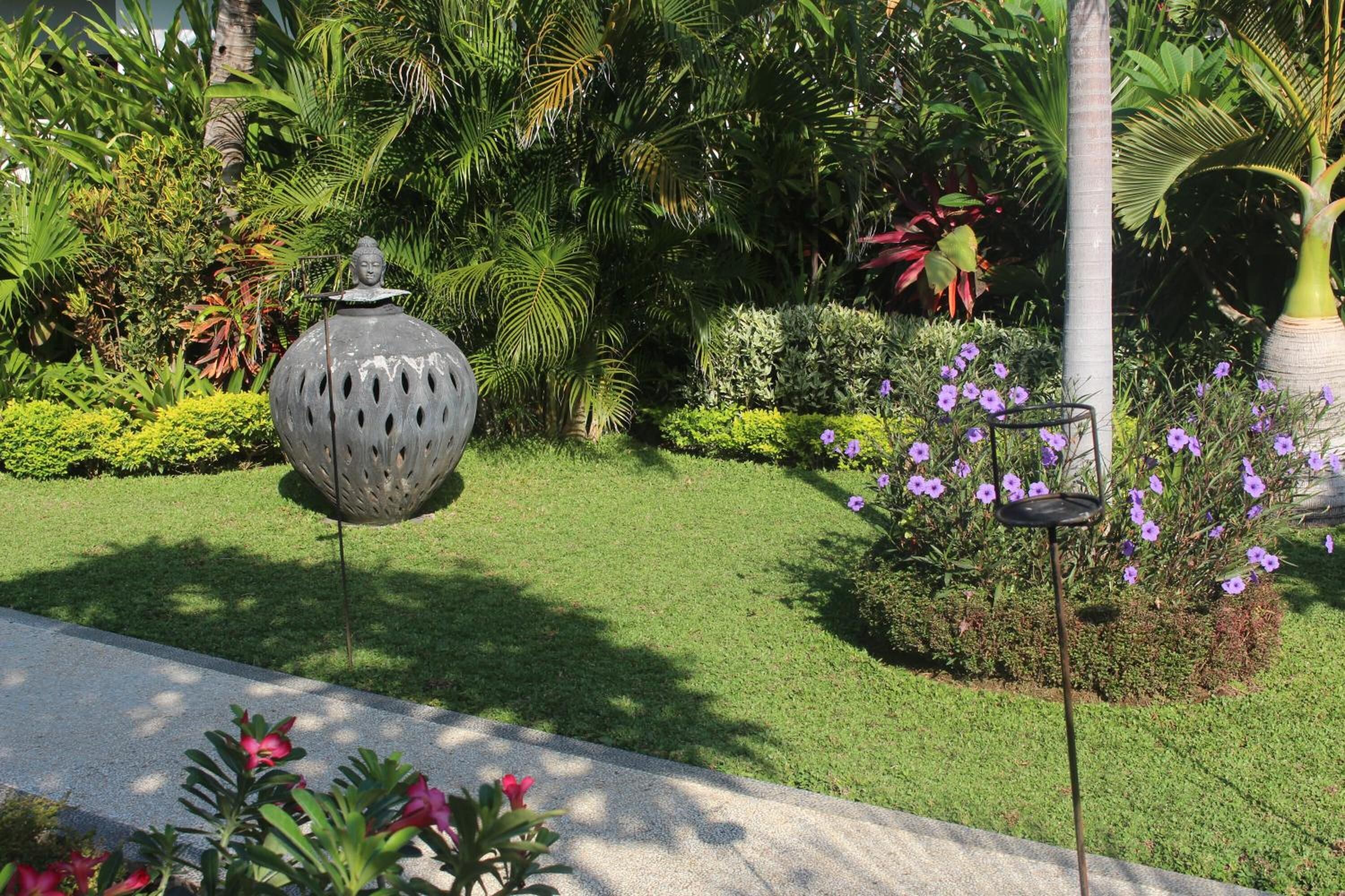 Un jardín con un florero metálico decorativo, una palmera alta y flores púrpuras.