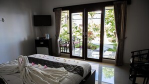 Une chambre d’hôtel avec un lit, un téléviseur fixé au mur, un petit bureau avec une bouteille d’eau, et une vue sur un jardin depuis les portes coulissantes.