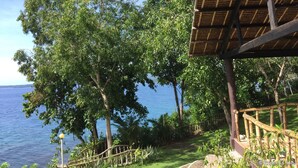A view of a beachside area with a wooden deck and railing, surrounded by lush greenery and trees. - Bohol Lahoy Dive Resort (Guindulman)
