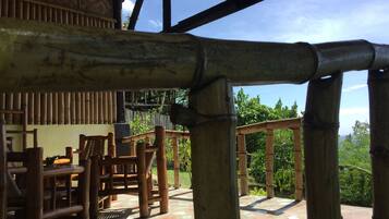 Un área de estar al aire libre con estructura de bambú, con sillas y una mesa, con vista a un jardín y un cielo despejado.