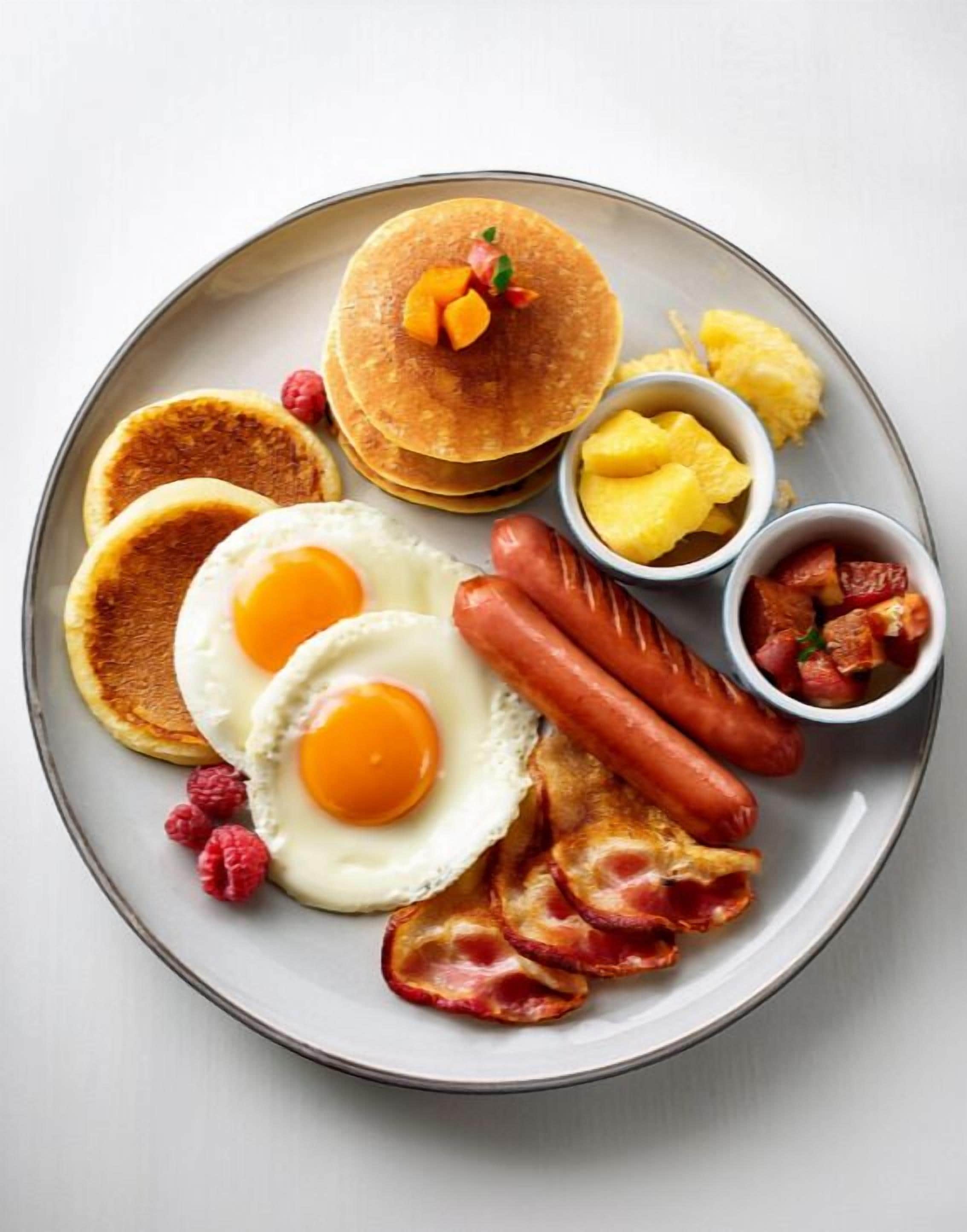Een ontbijtplaat met pannenkoeken, eieren, worstjes, spek, frambozen en fruit.