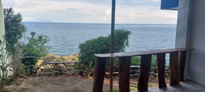 A wooden table and bench by a window overlooking a calm sea with distant mountains.