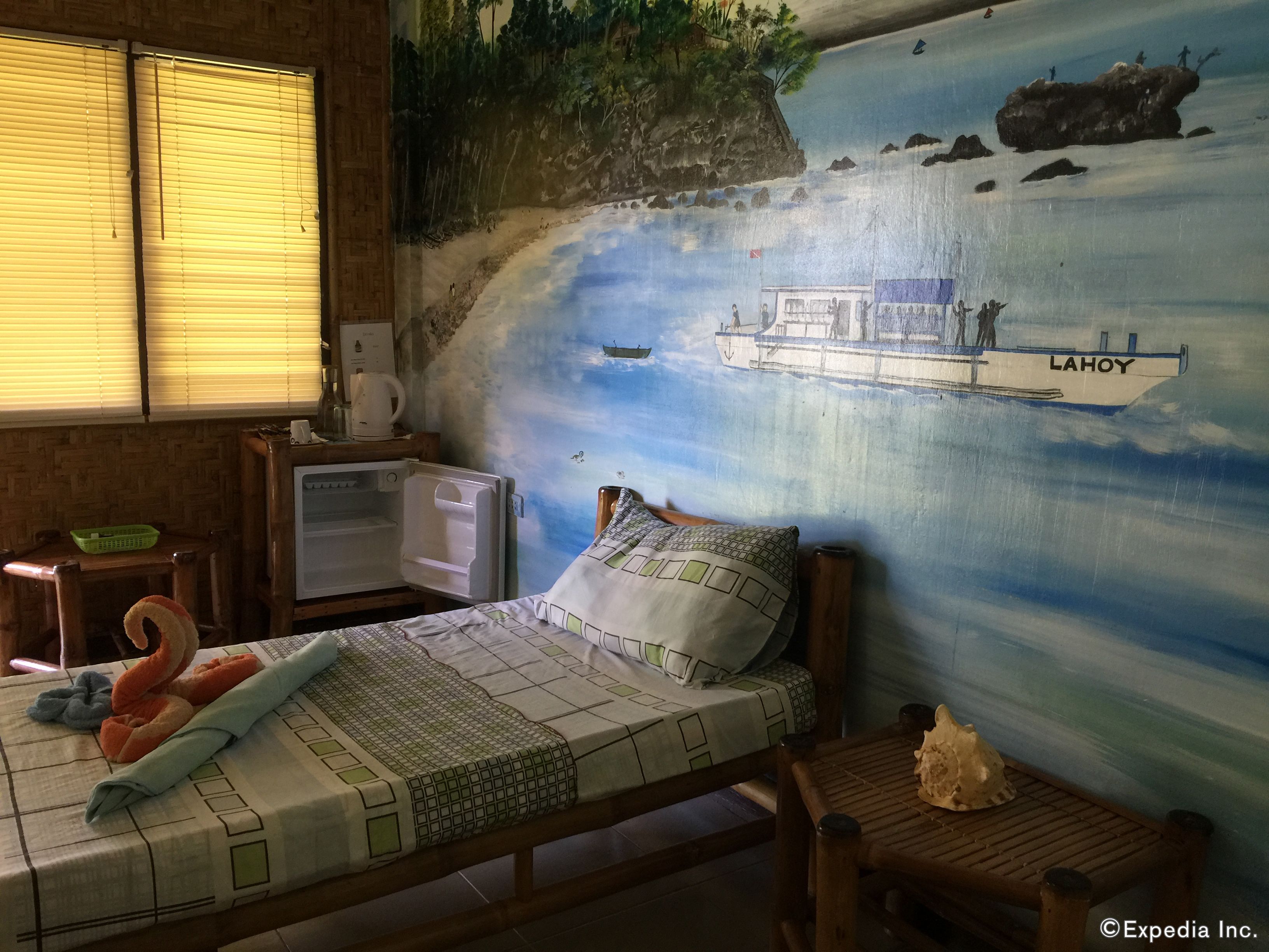 Una habitación con cama, una mesita, un refrigerador y un mural de un barco en el agua.