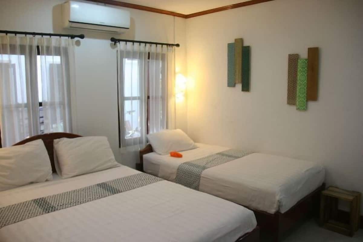 A hotel room with two beds, a nightstand, a small table, an air conditioning unit, and a window with curtains.