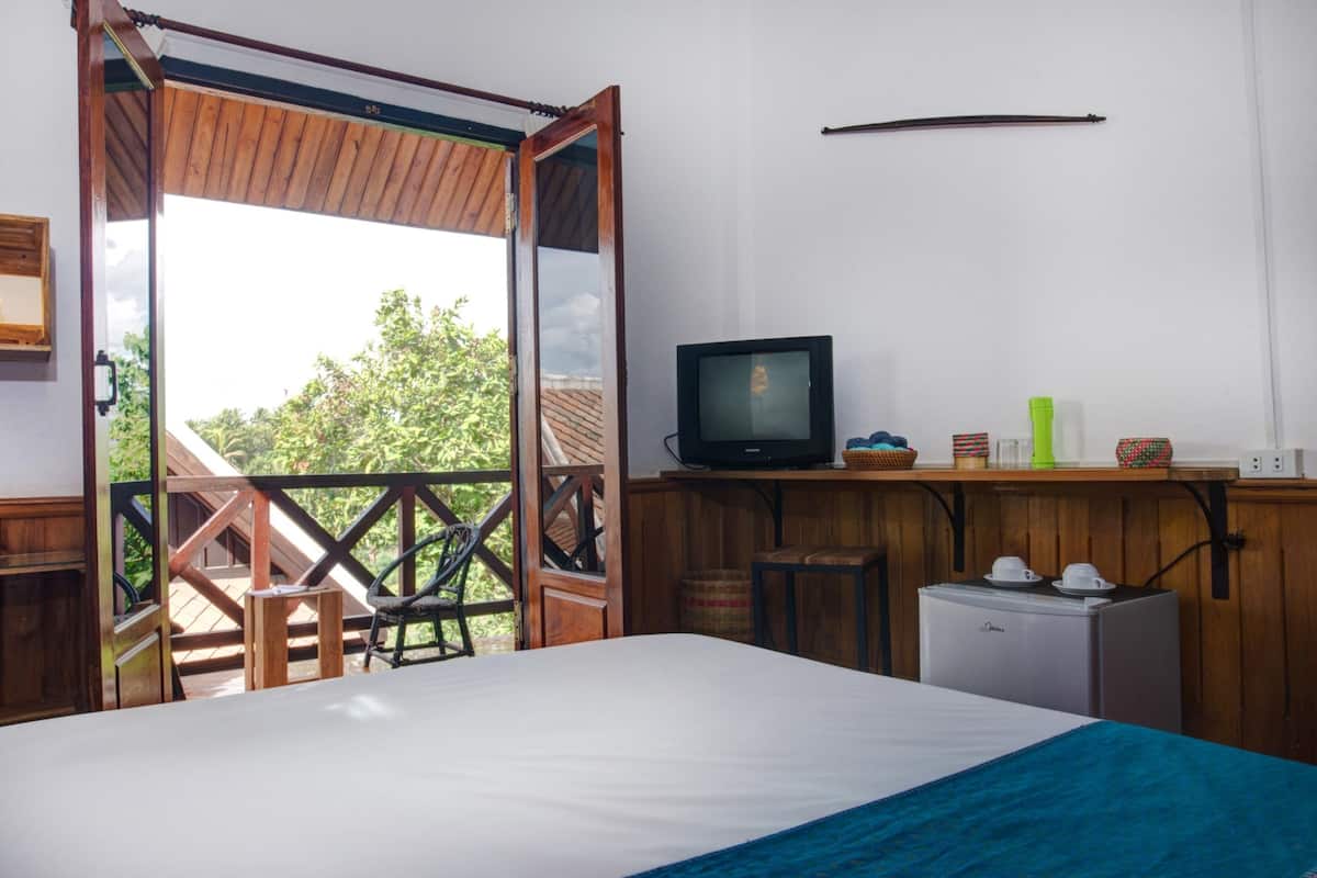 A bedroom with a bed, a television, a small refrigerator, and a balcony with a wooden railing.