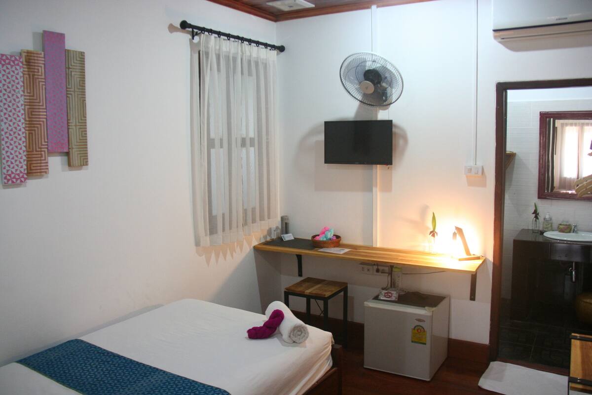 A bedroom with a bed, a desk with a lamp, a small refrigerator, a mirror, and a bathroom with a sink.