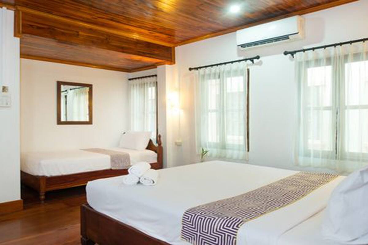A hotel room with a wooden ceiling, two beds, a large window with curtains, and an air conditioning unit.