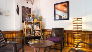 A cozy reading nook with a wooden bookshelf, a round table with a purple cushion, and a tall floor lamp. - Cold River (Luang Prabang)