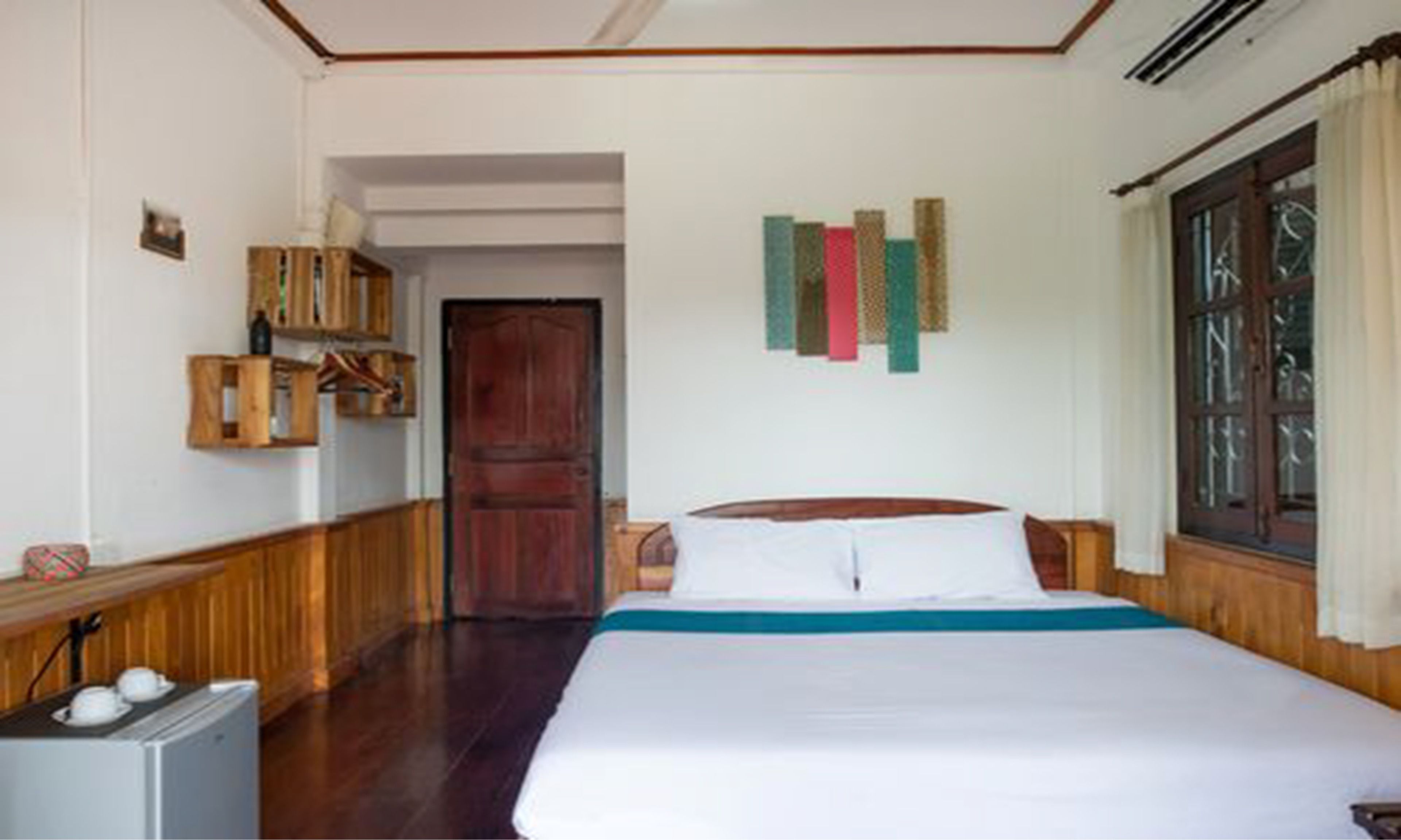 Una habitación de hotel con cama, un armario de madera, un refrigerador pequeño y una ventana con cortinas.