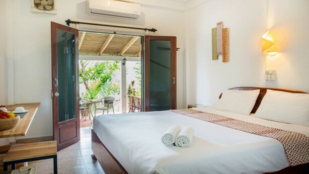 A hotel room with a bed, a small table with a basket, a chair, and a view of the outdoors through an open door. - Cold River (Luang Prabang)