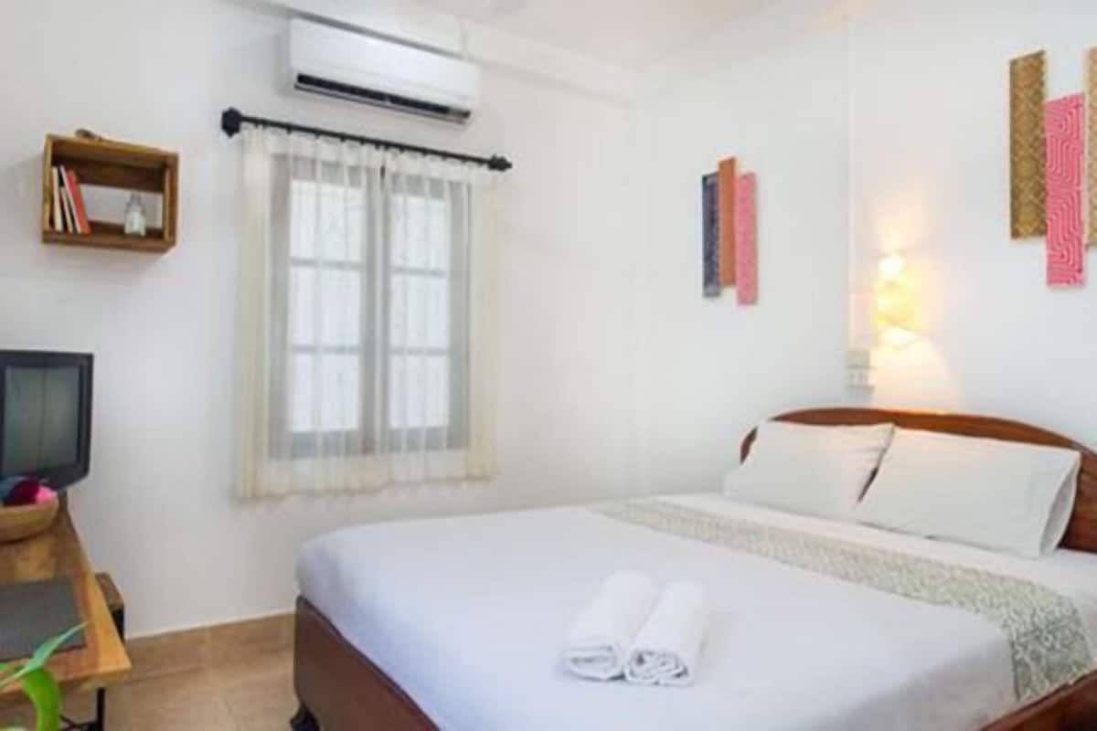 A hotel room with a bed, a TV, a shelf with books, an air conditioner, and a window with curtains.